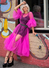 A woman models a vibrant magenta pink tulle midi dress with a deep purple mesh bodice, a black studded belt, and dramatic puff sleeves, posing against a brick wall with graffiti.
