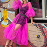 A woman models a vibrant magenta pink tulle midi dress with a deep purple mesh bodice, a black studded belt, and dramatic puff sleeves, posing against a brick wall with graffiti.