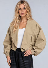 A blonde model wearing a cropped, oversized tan utility jacket with elasticized cuffs, styled over a white tank top and dark plaid pants, against a solid light gray background.