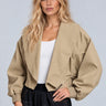 A blonde model wearing a cropped, oversized tan utility jacket with elasticized cuffs, styled over a white tank top and dark plaid pants, against a solid light gray background.