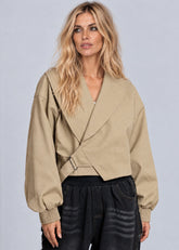 A woman modeling a cropped beige wrap-style jacket with a wrap front, belted detail, and ribbed cuffs, paired with dark wide-leg jeans against a light gray studio background.