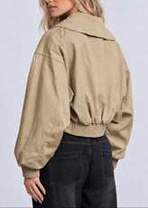 A woman shown from the back wearing a cropped, oversized tan utility jacket with a wide collar and elasticated hem, paired with dark jeans, against a light gray studio background.