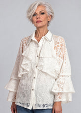 A woman with short gray hair wearing a white lace blouse with ruffles and sheer sleeves, paired with blue jeans, against a solid gray studio background.