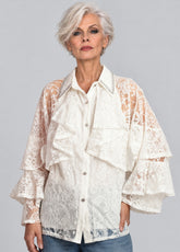 A woman with short gray hair wearing a cream-colored lace blouse with ruffles and sheer patterned sleeves, paired with blue jeans, against a solid gray studio background.