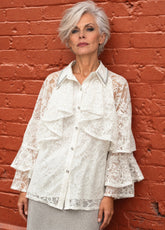 A woman with short gray hair wearing a white lace ruffled blouse, standing against a textured red brick wall.