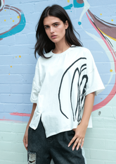 Woman wearing a white t-shirt with black abstract design against a graffiti-covered wall.