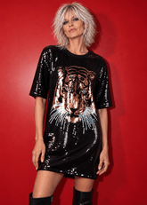 A model with short silver hair wears a black sequined t-shirt dress featuring a large sequined tiger face graphic on the front, paired with black leather knee-high boots, set against a solid red background.